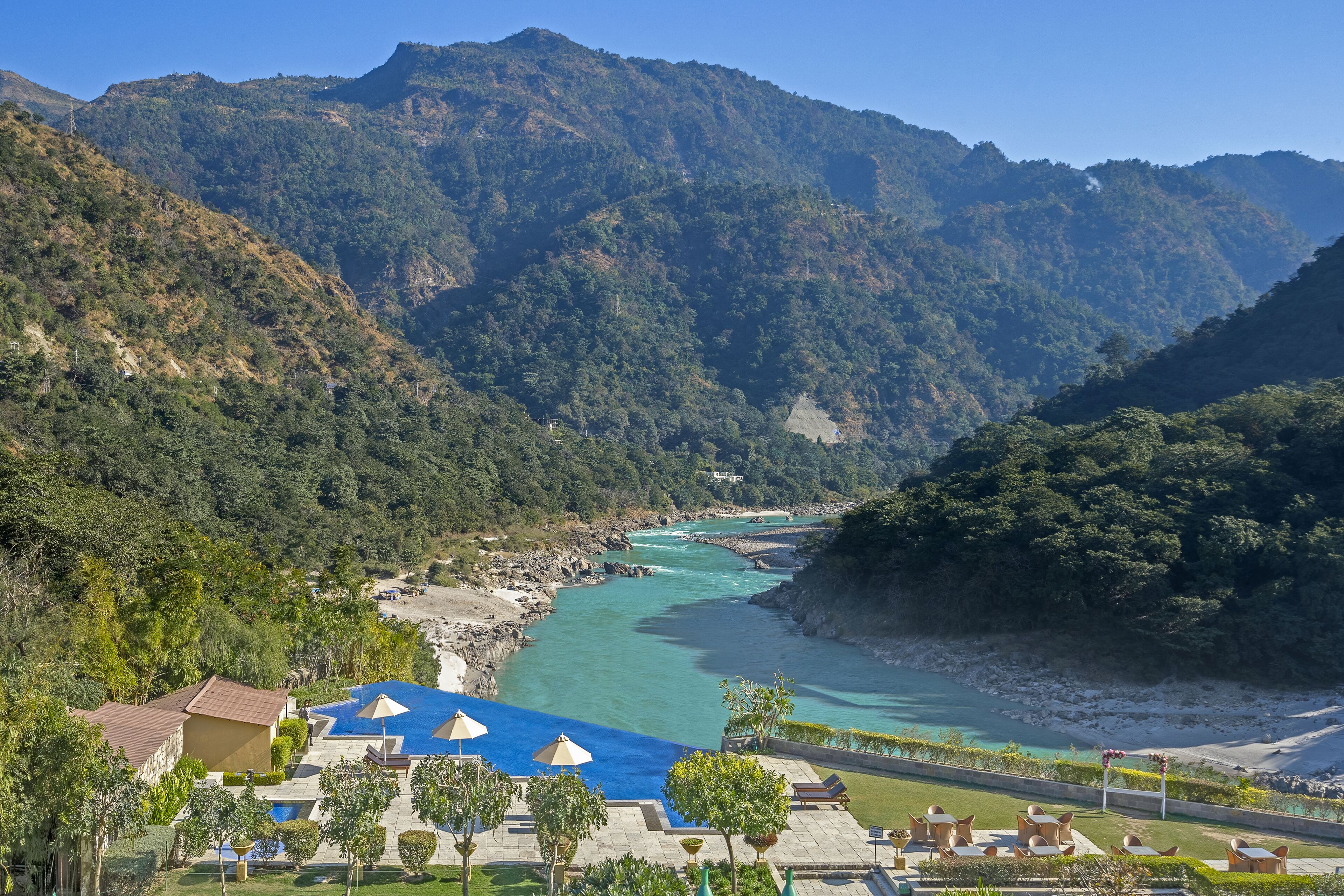 A resort with a swimming pool, lounge chairs, and a view of a river and mountains.