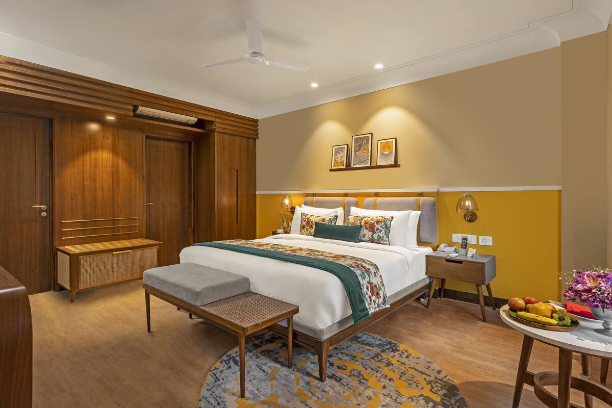 Quarto de hotel moderno com uma cama grande, mesas de cabeceira, um espaço de estar pequeno e uma mesa de jantar com frutas.