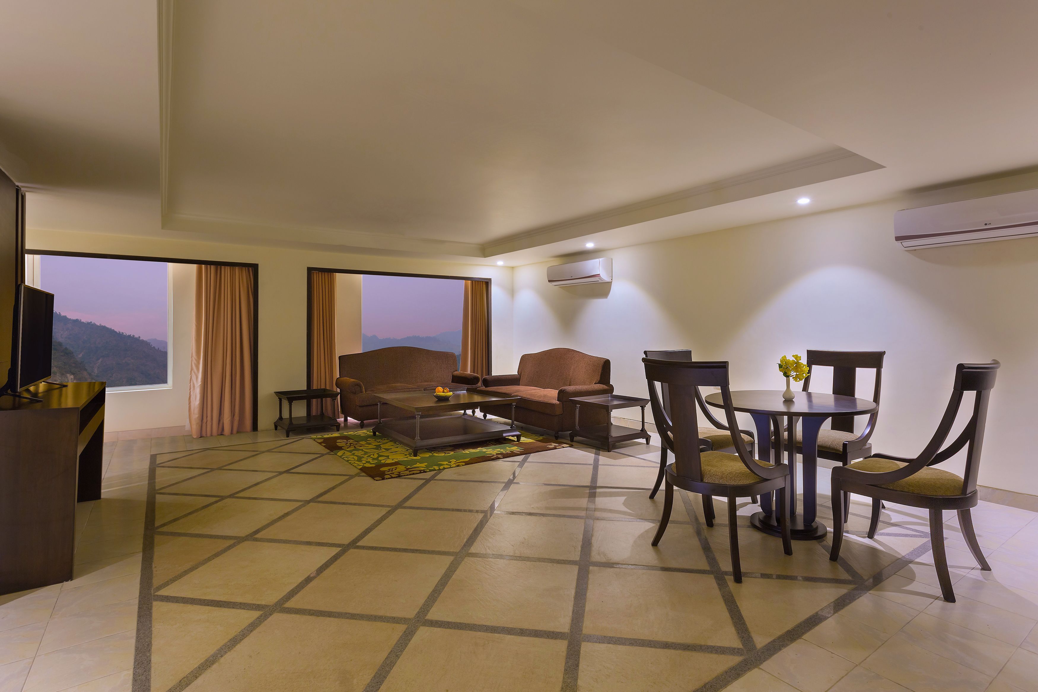 A spacious living area with a sofa set, a coffee table, and a dining table with chairs. There are large windows offering a view of the mountains.