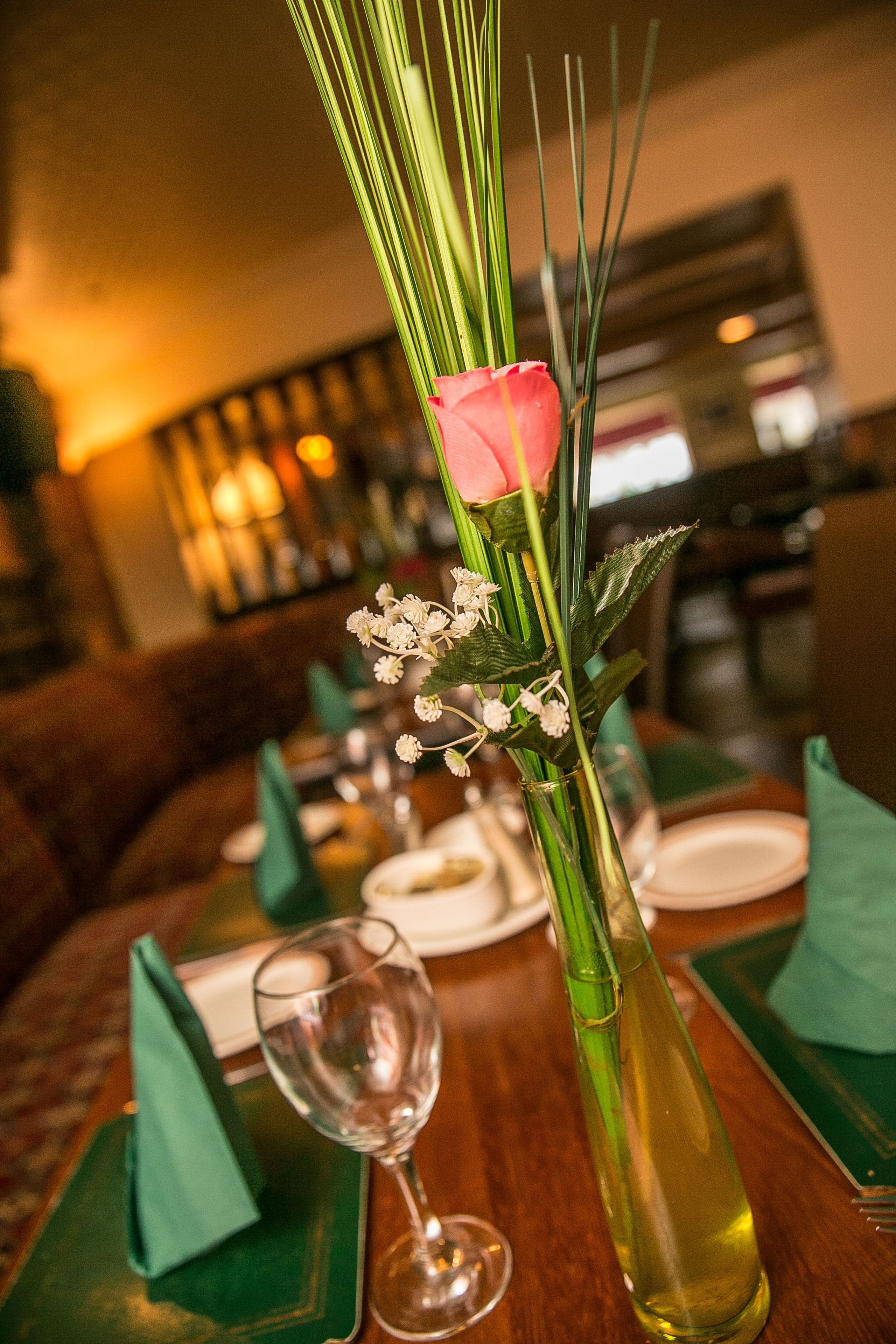 A dining table with a vase containing a pink rose, green foliage, and small white flowers.