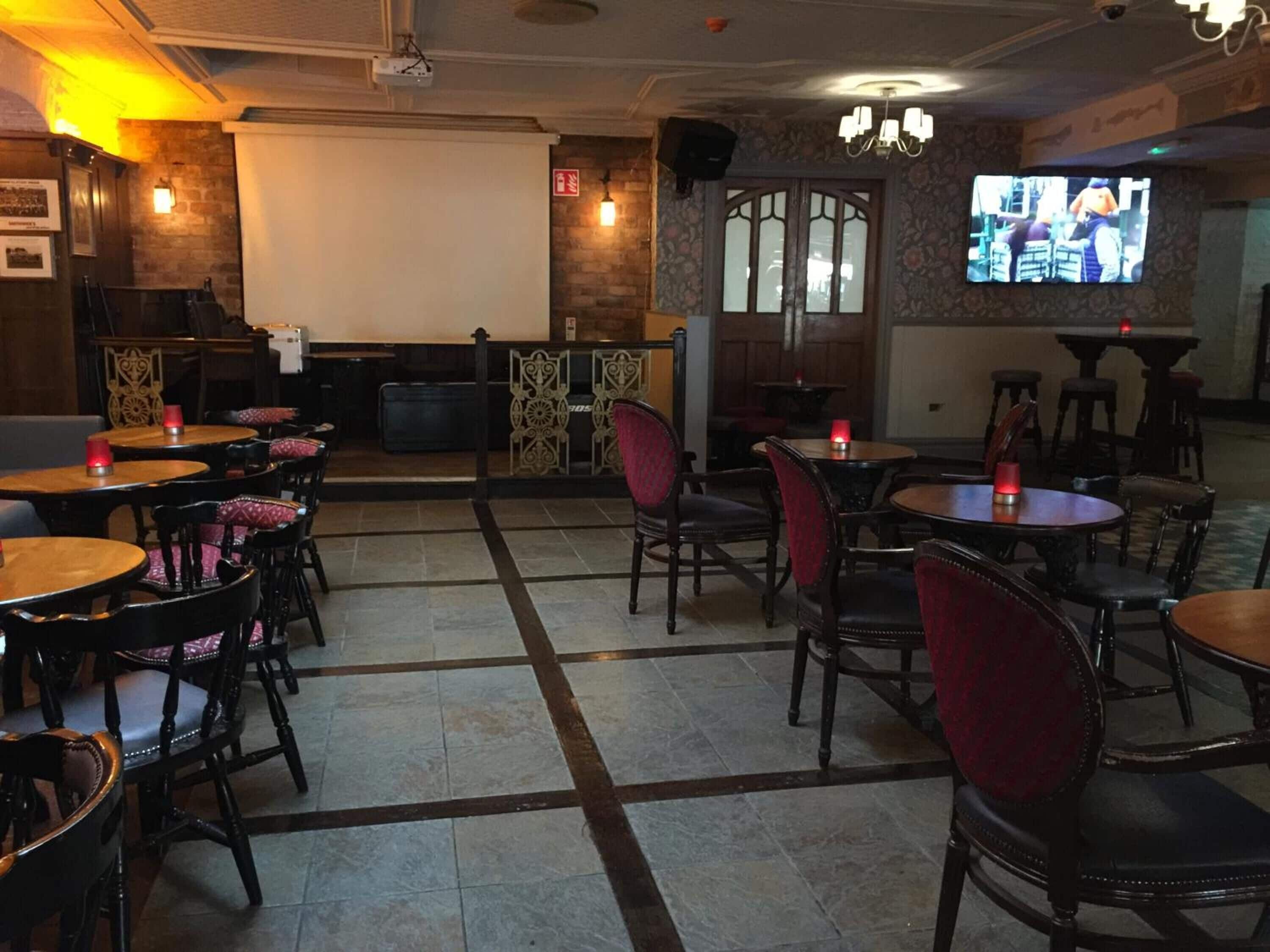 A restaurant with wooden tables and chairs, a bar area, and a television screen displaying a show.