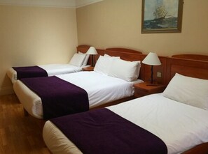 A hotel room with two single beds, each with a wooden headboard, white bedding, and a purple throw blanket. There is a nightstand with a lamp on each bed, and a painting of a ship on the wall.