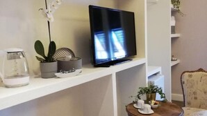 Una sala de estar con una televisión sobre un estante blanco, una mesita con tazas, un sillón y una planta.