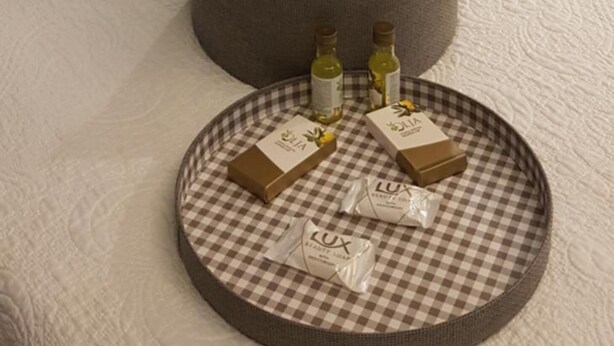 Una cama bien hecha con sábanas blancas y una mesita de noche gris trenzada, con toallas dobladas y una bandeja con artículos de tocador.