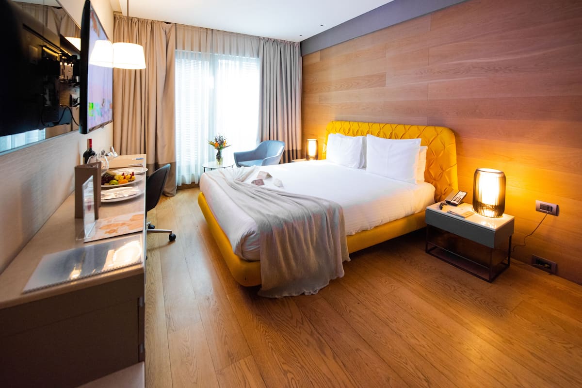 A hotel room with a bed, bedside table, lamp, TV, and a desk with a chair and a fruit basket.