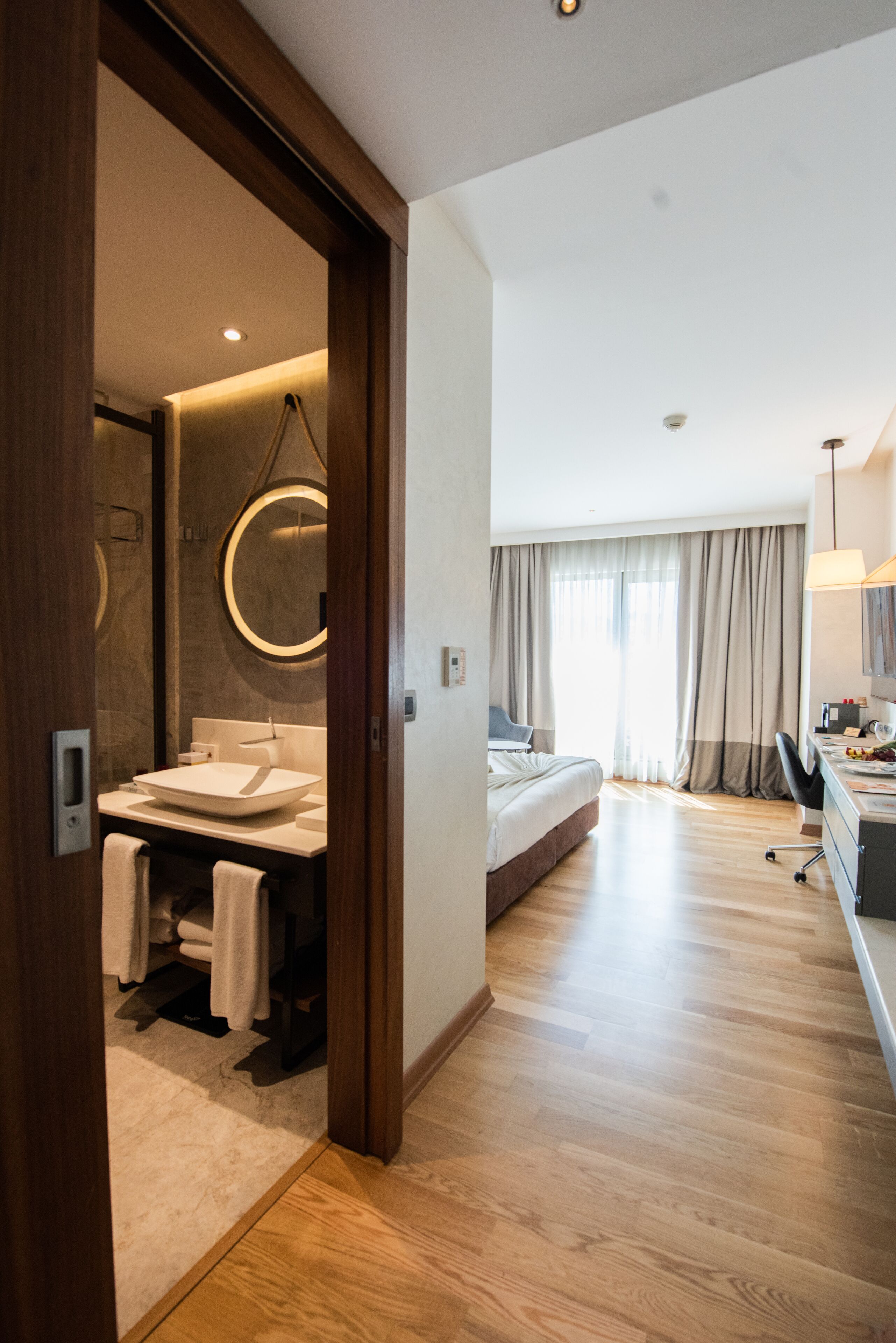 A modern hotel room with a wooden floor, a large bed, a desk with a chair, a round mirror, and a bathroom with a sink and towel rack.