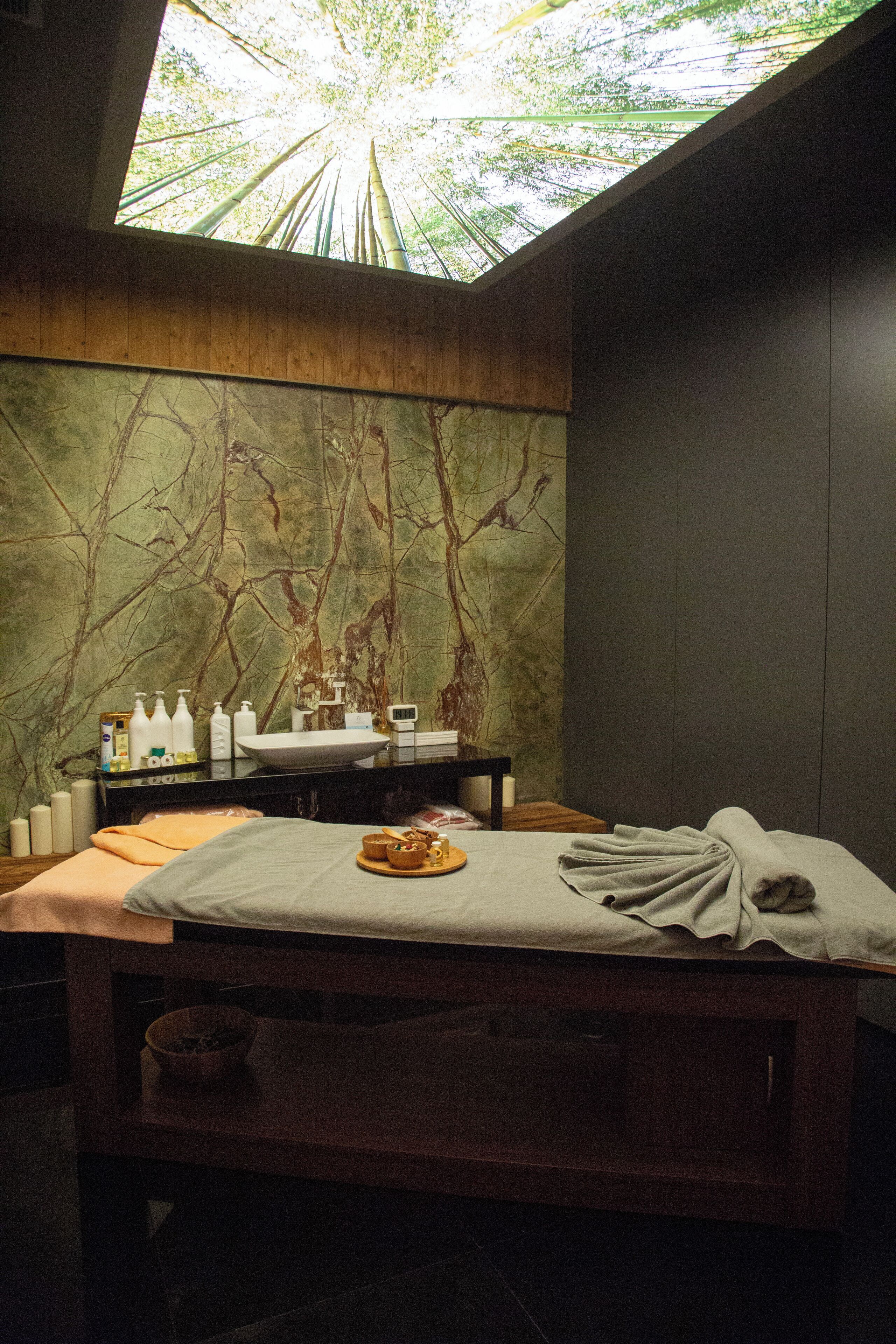 Ett spa-rum med ett stort takfönster, en träbänk, ett massagebord och en vägg dekorerad med en väggmålning som föreställer en trägren.
