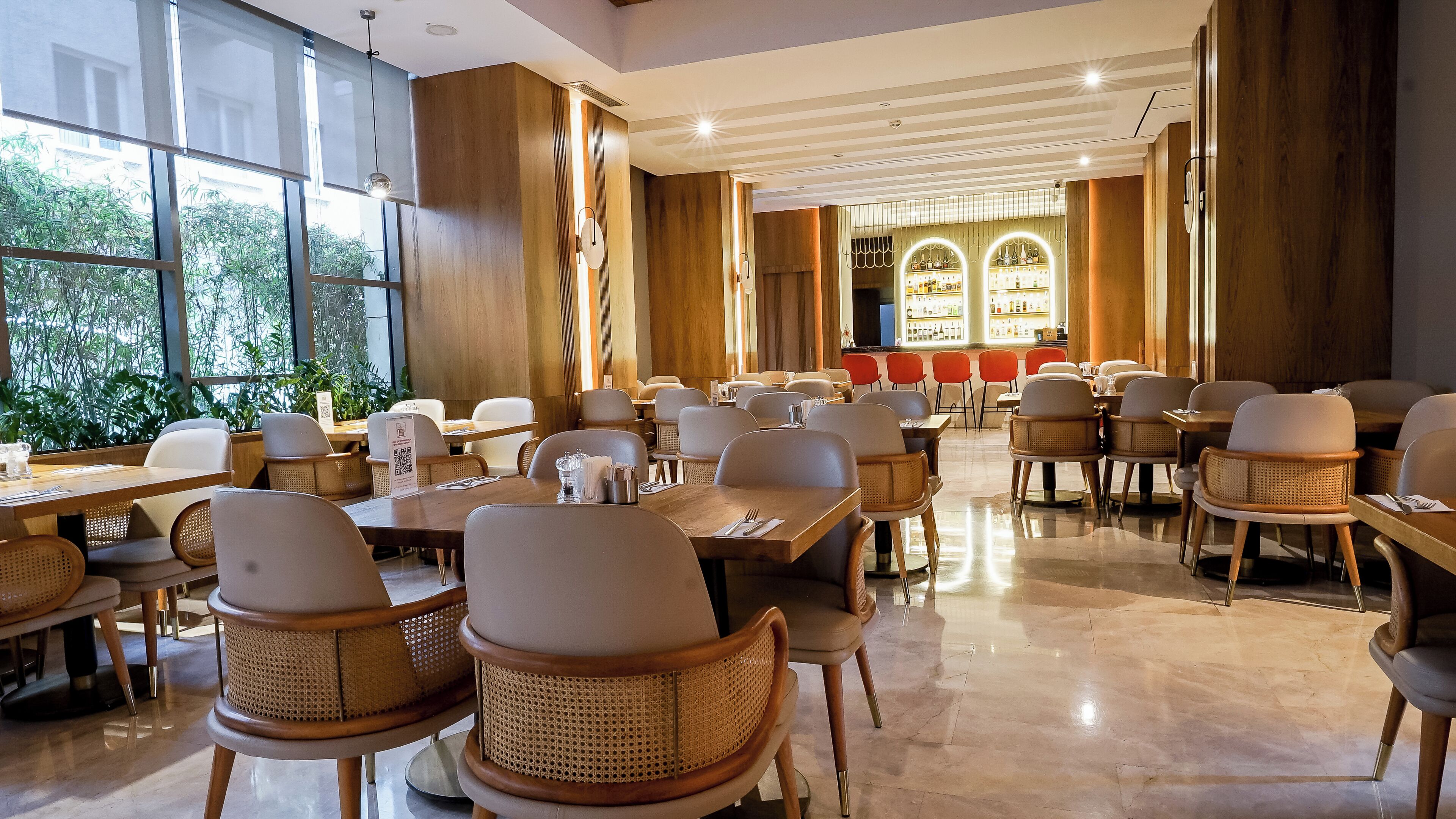 A spacious dining area with high tables and wicker chairs, large windows, and a bar area with illuminated shelves.