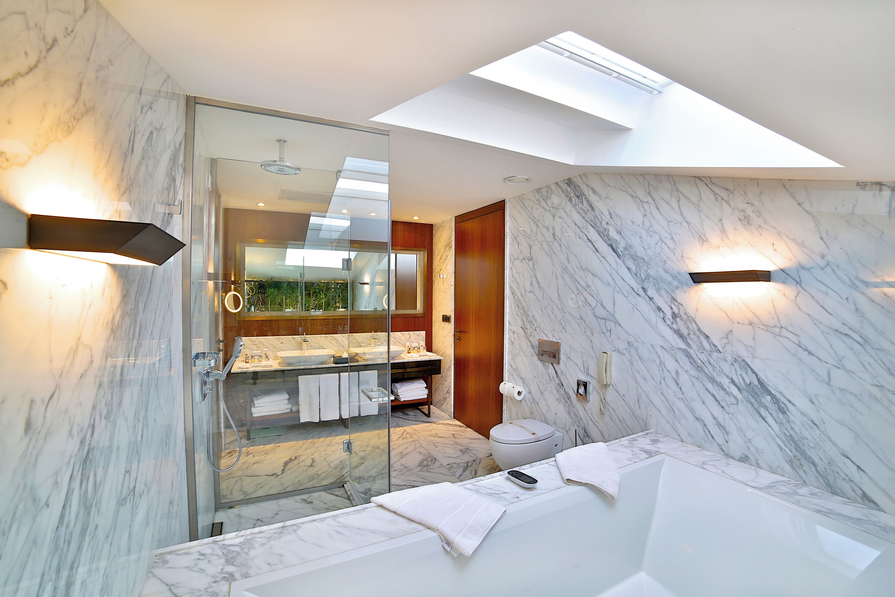 A modern bathroom with a skylight, marble walls, a glass shower enclosure, and a white sink.