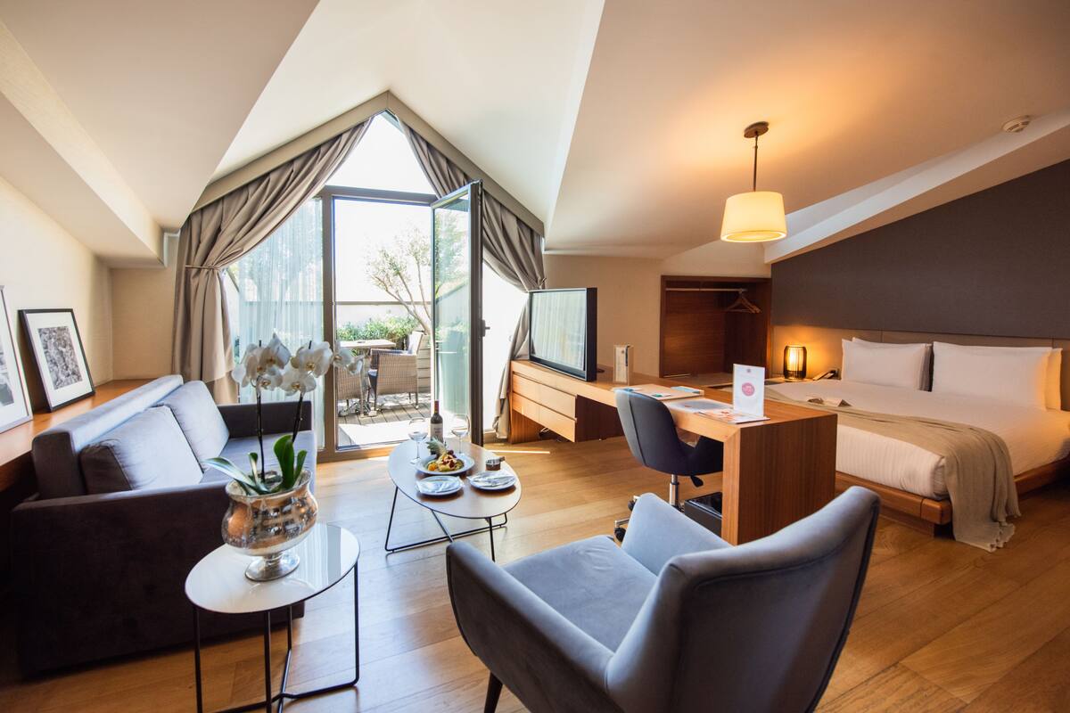 A modern hotel room with a loft-style ceiling, a cozy seating area, a dining table, a workspace, and a large bed.