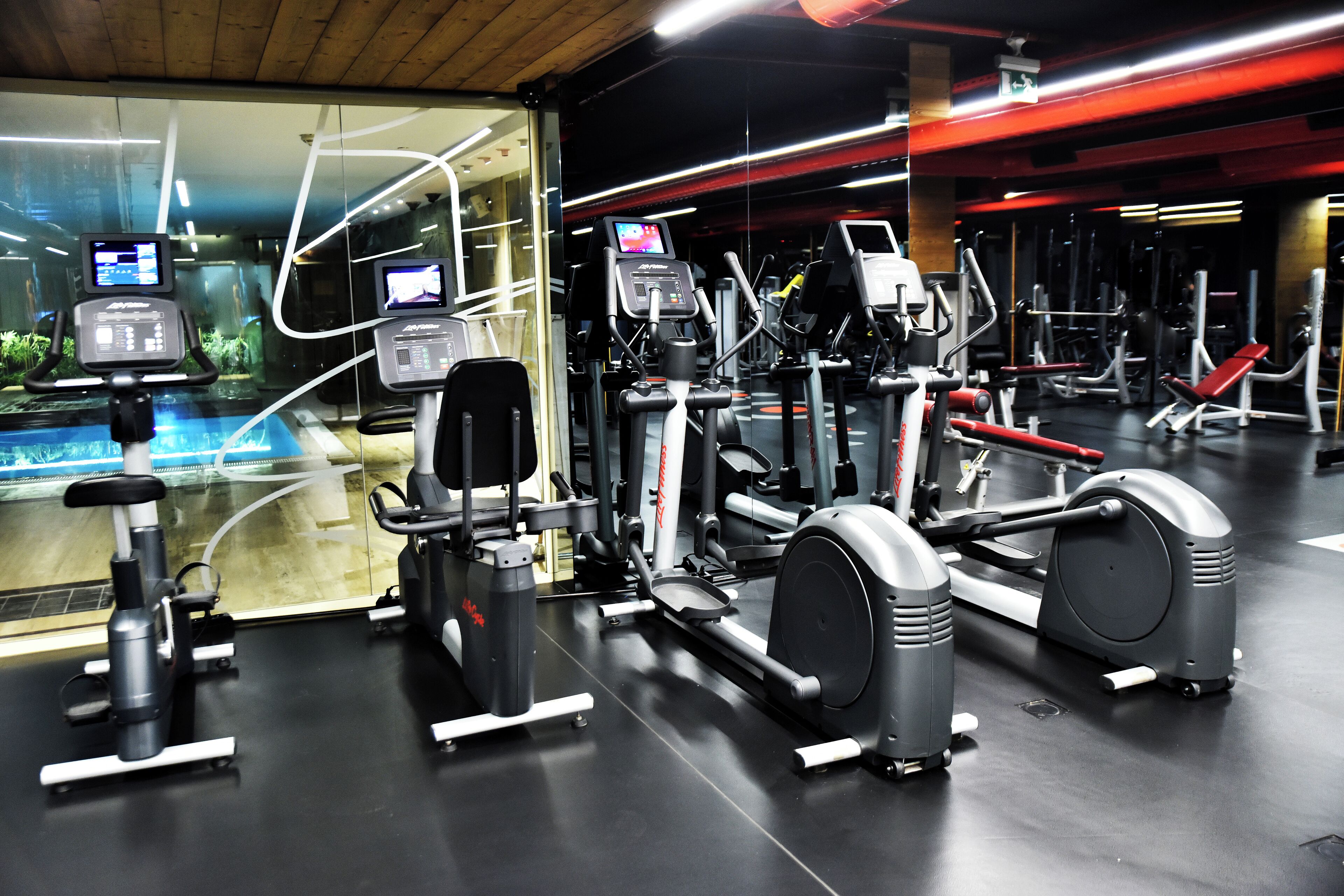 Ein Fitnessstudio mit verschiedenen Trainingsgeräten, einem großen Spiegel und einem Fenster mit Blick auf ein Gebäude.