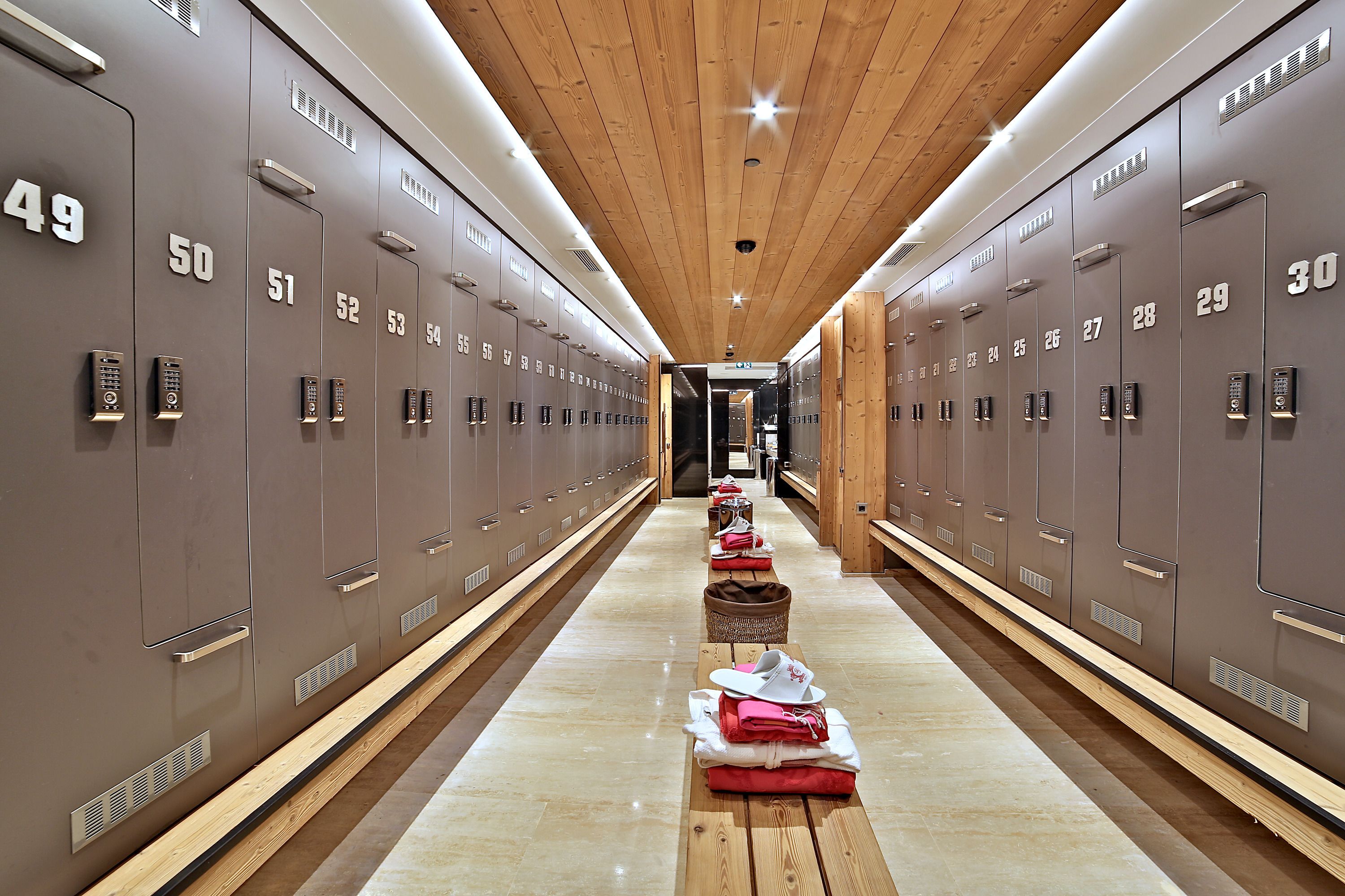 A hotel corridor with lockers numbered 30 to 54, wooden flooring, and a central bench with folded towels and hats.