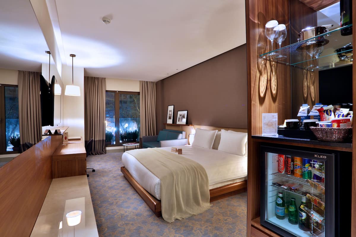 A hotel room with a large bed, a seating area, a minibar, and a view of the outdoors.