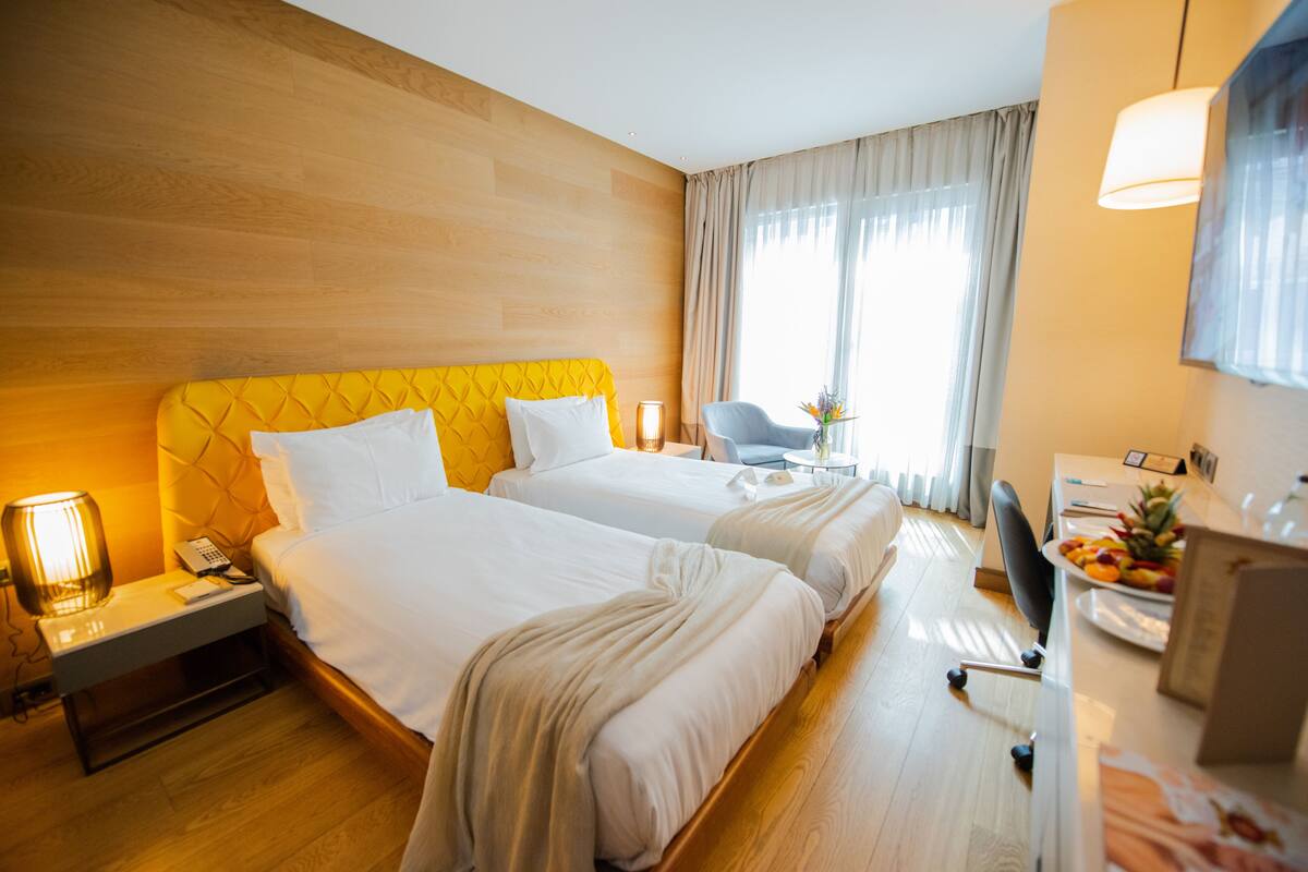 A hotel room with two beds, a desk with a chair, a lamp, and a fruit bowl.