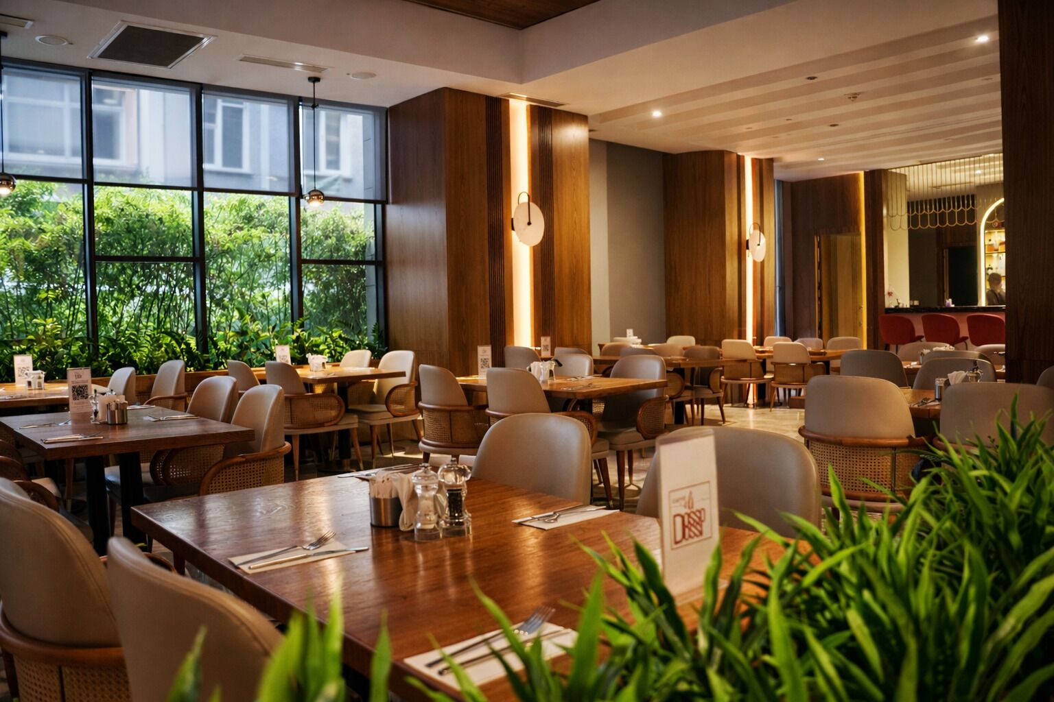 Un restaurant doté de nombreuses tables et chaises, de grandes fenêtres offrant une vue sur la verdure et d’un aménagement intérieur moderne.