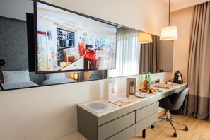 A modern hotel room with a large mirror reflecting the room's interior, a desk with a laptop, a coffee maker, and a fruit bowl.