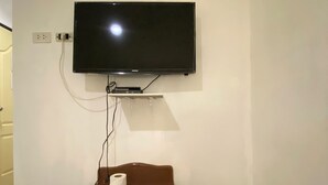 Une pièce équipée d’un téléviseur à écran plat fixé au mur, d’une table en bois sur laquelle se trouve un rouleau de papier toilette, et d’une boîte de rangement jaune.