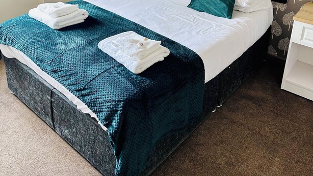 A neatly made bed with a teal bedspread and pillows, a white headboard, and a small bedside table with a lamp. - The Minworth Club & Lodge  (Sutton Coldfield)