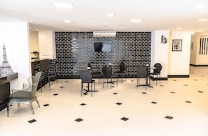 A hotel lobby with a black brick wall, a flat-screen TV mounted above a reception desk, and several chairs and tables arranged for guests.