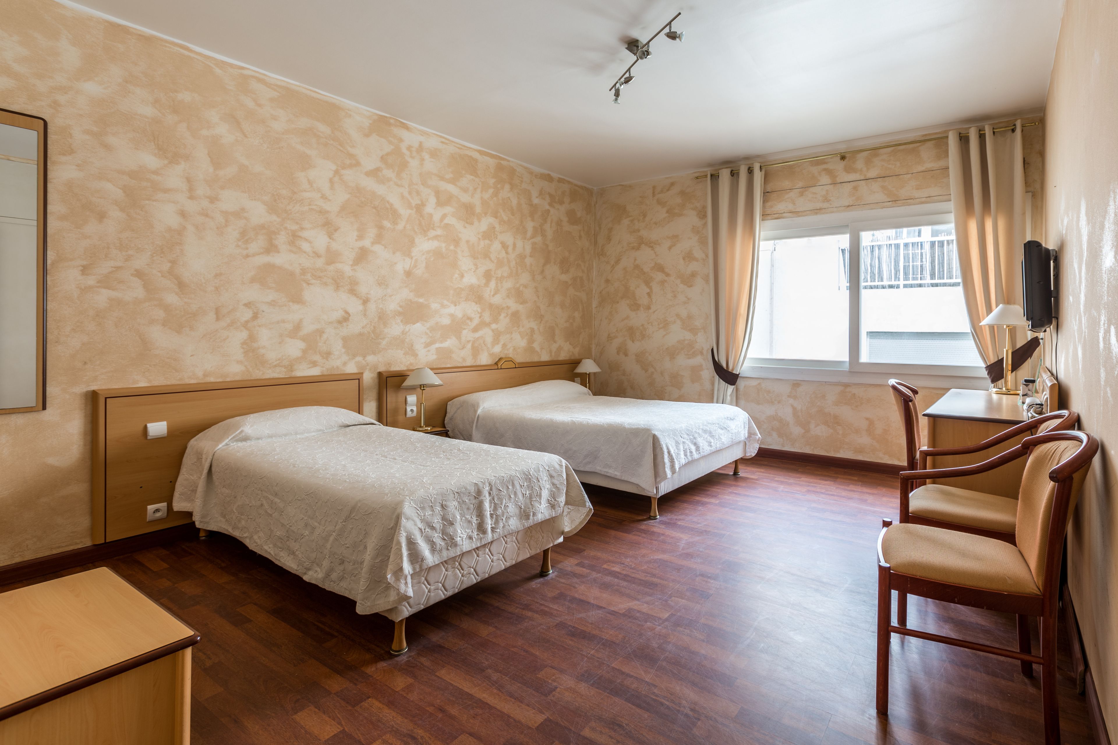 Une chambre d’hôtel avec deux lits simples, un bureau en bois, une chaise, un téléviseur fixé au mur et une fenêtre avec des rideaux.