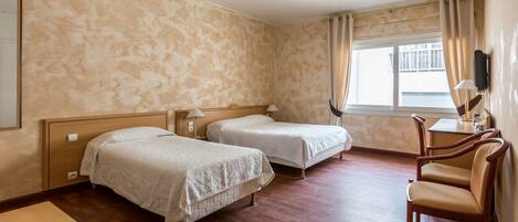 A hotel room with two single beds, a wooden desk, a chair, a TV mounted on the wall, and a window with curtains.