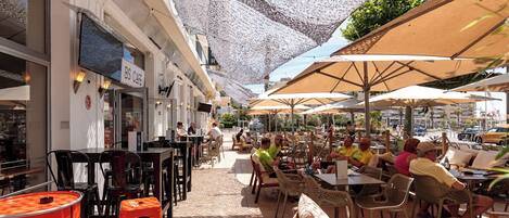 Um café ao ar livre com mesas e cadeiras, um letreiro com 'BS CAFE' e um chão de azulejos.