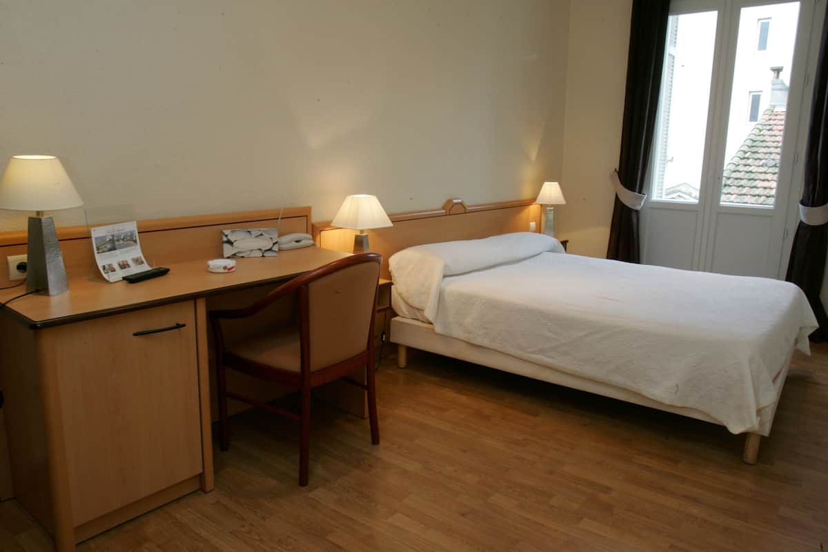 A hotel room with a wooden desk, two bedside lamps, a bed with white linens, and a window with curtains.