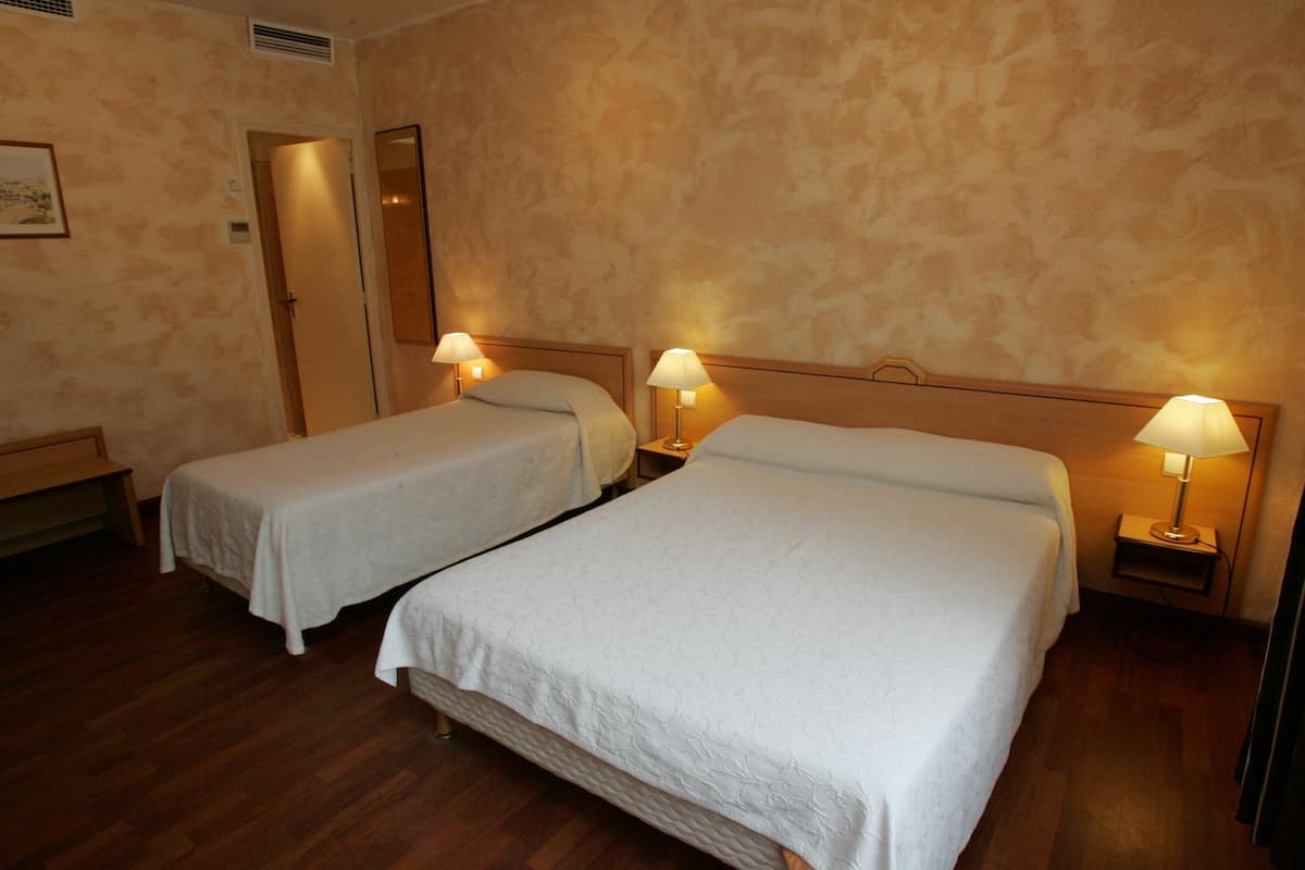 A hotel room with two single beds, each with a bedside table and lamp, wooden flooring, and a door leading to another room.