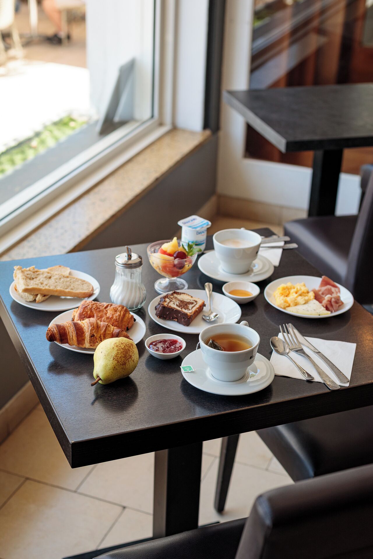 Een ontbijttafel met croissants, fruit, jam, roerei, ham en een kop thee.