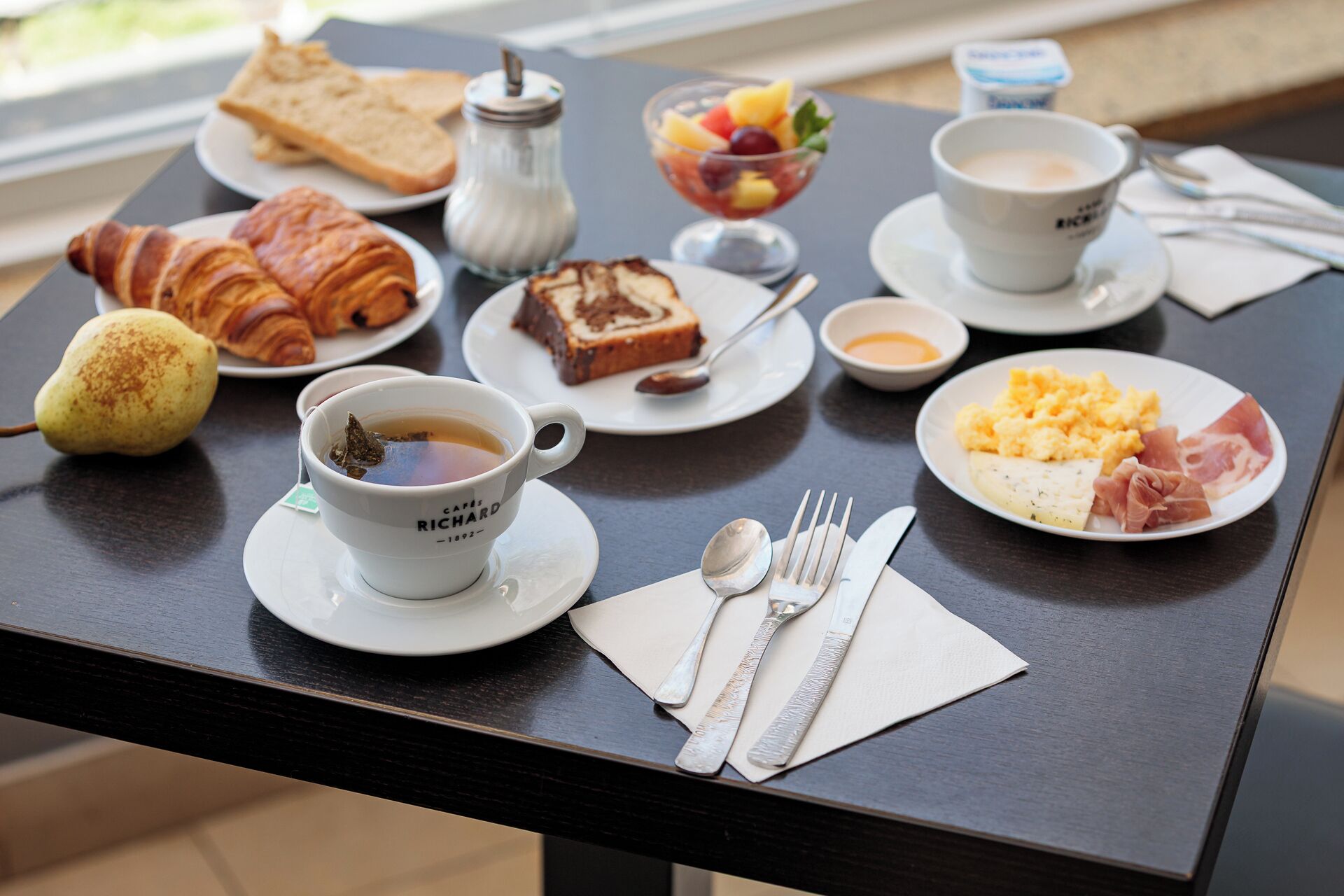 Una mesa de desayuno con croissants, tostadas, fruta, huevos revueltos, jamón y café.
