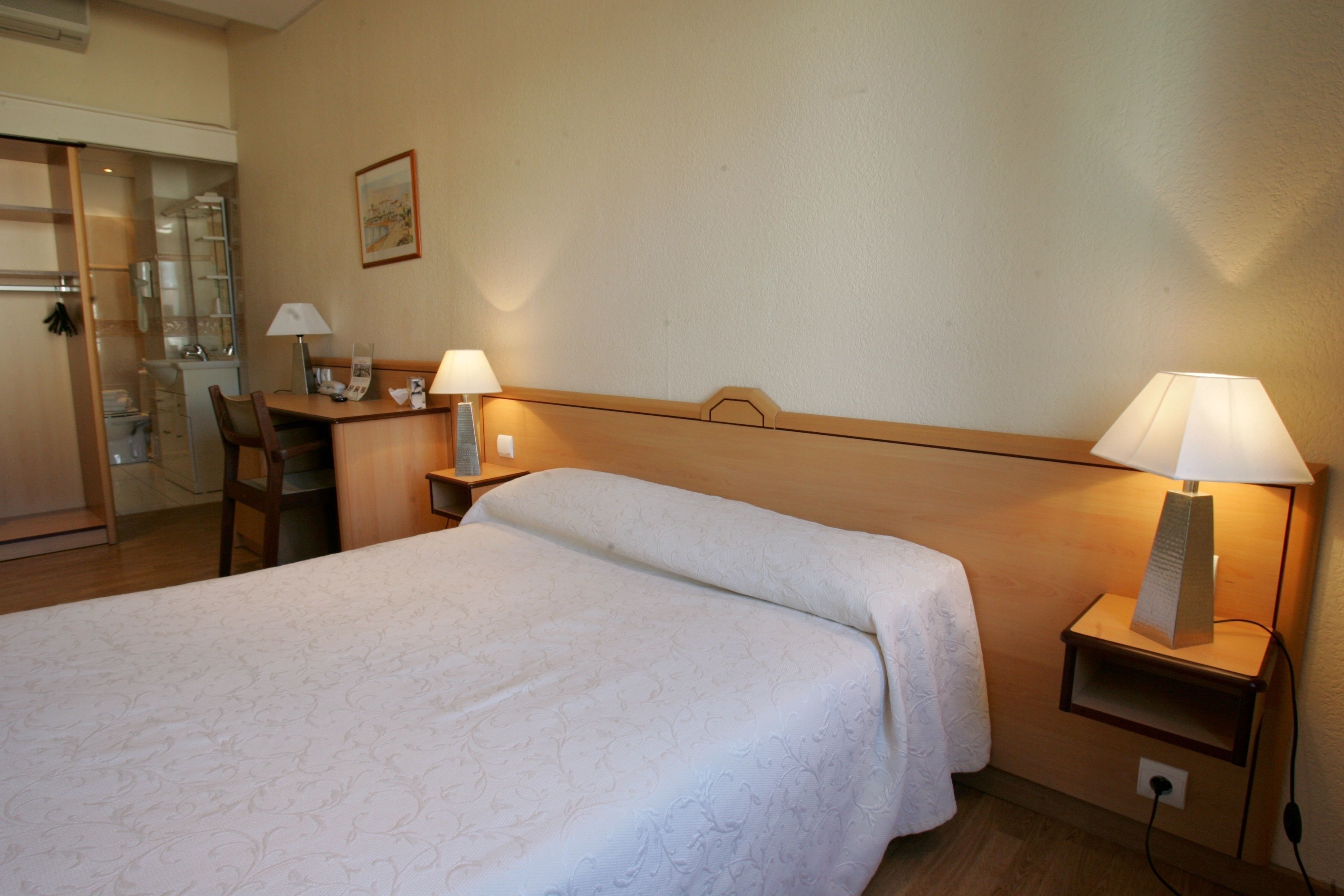 Une chambre d’hôtel comprenant un lit, un bureau, deux lampes de chevet, une table de chevet et une petite étagère.
