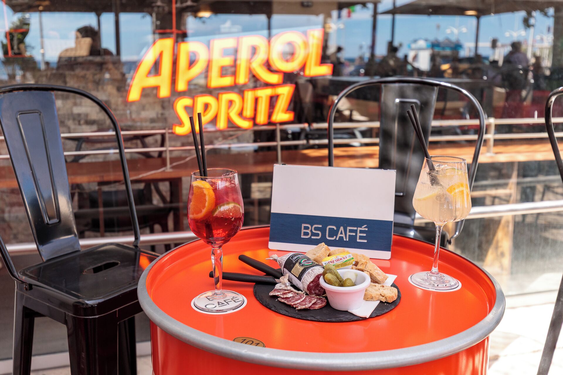 Een tafel met een bord waarop 'BS CAFÉ' staat, en twee drankjes, waarvan één met een oranjeschijfje en de ander met een citroenschijfje.