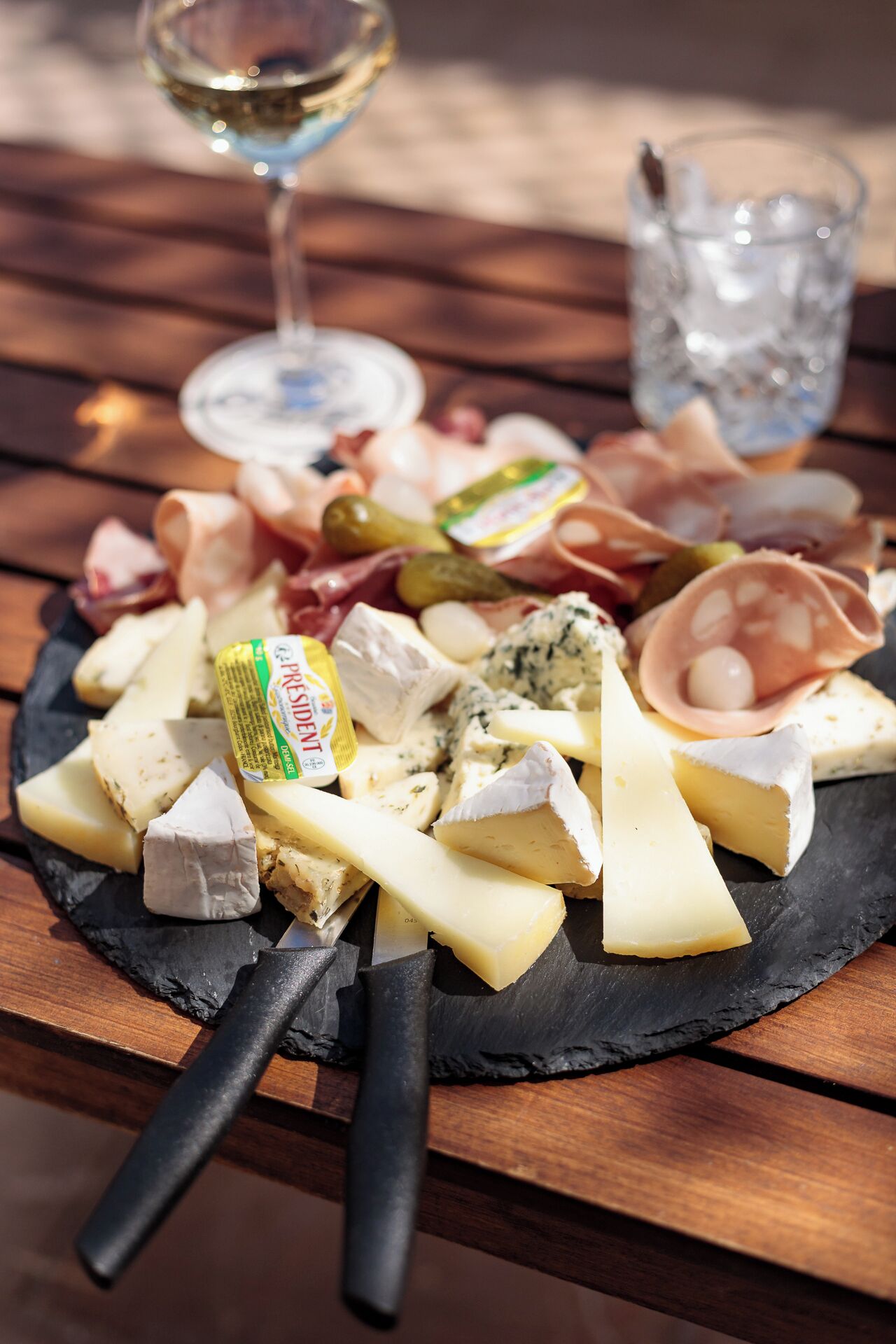 A charcuterie board with various cheeses, meats, and pickles on a slate board, accompanied by a glass of white wine and a glass of water.