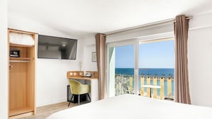 Habitación de hotel con cama, escritorio con silla, televisor, teléfono y balcón con vista al mar.