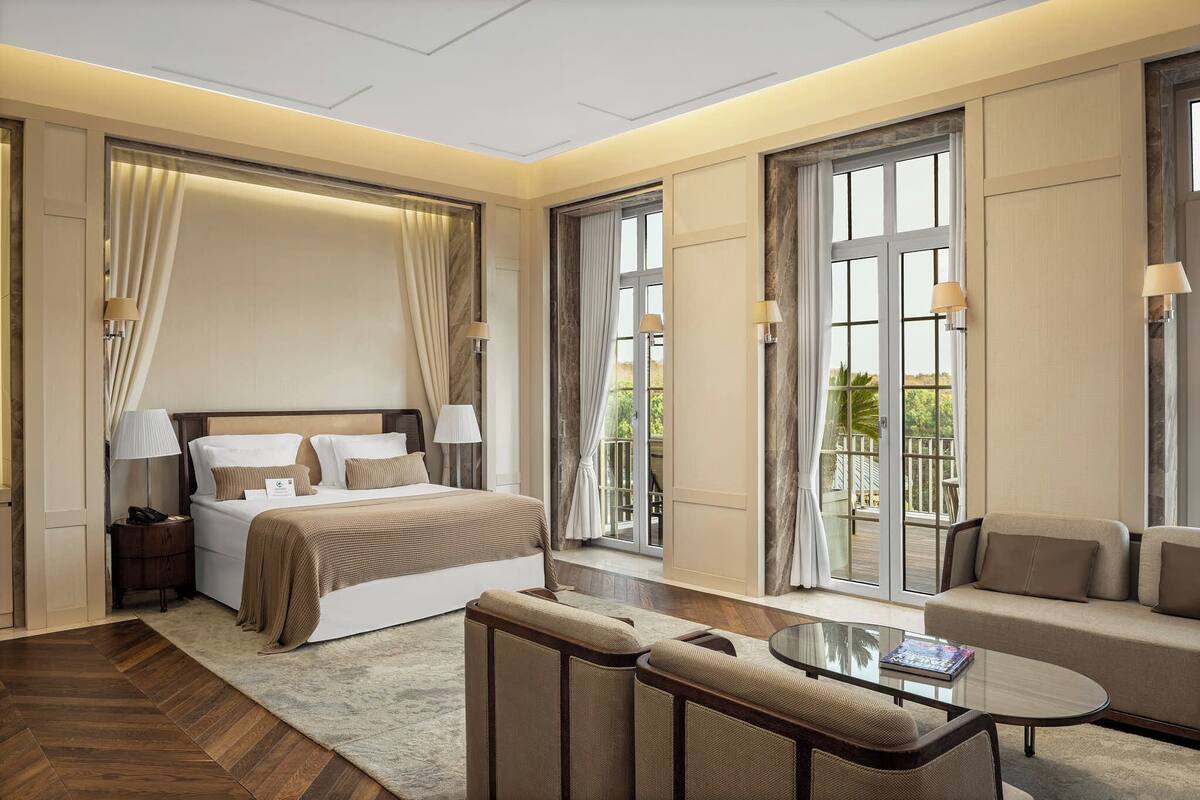 A modern hotel room with a large bed, a sofa, a glass coffee table, and a balcony with a view.