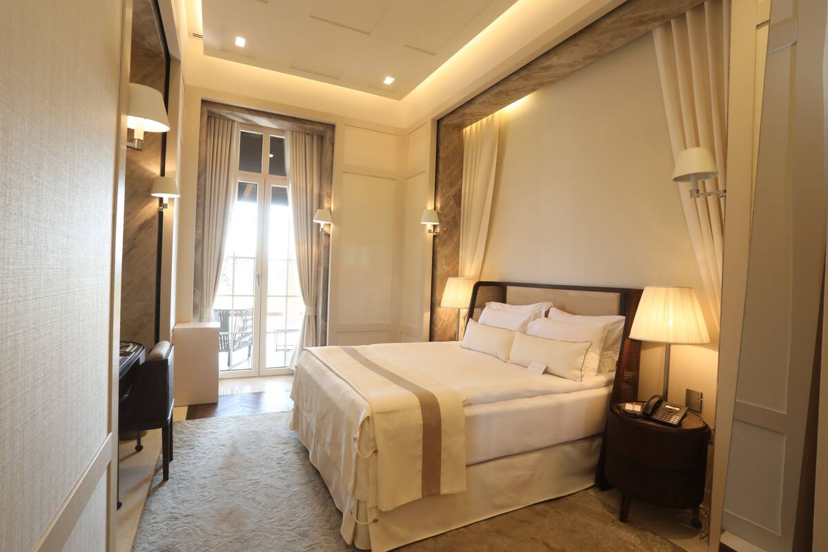 A hotel room with a large bed, bedside lamps, a desk with a chair, and a view of the outdoors through a window.