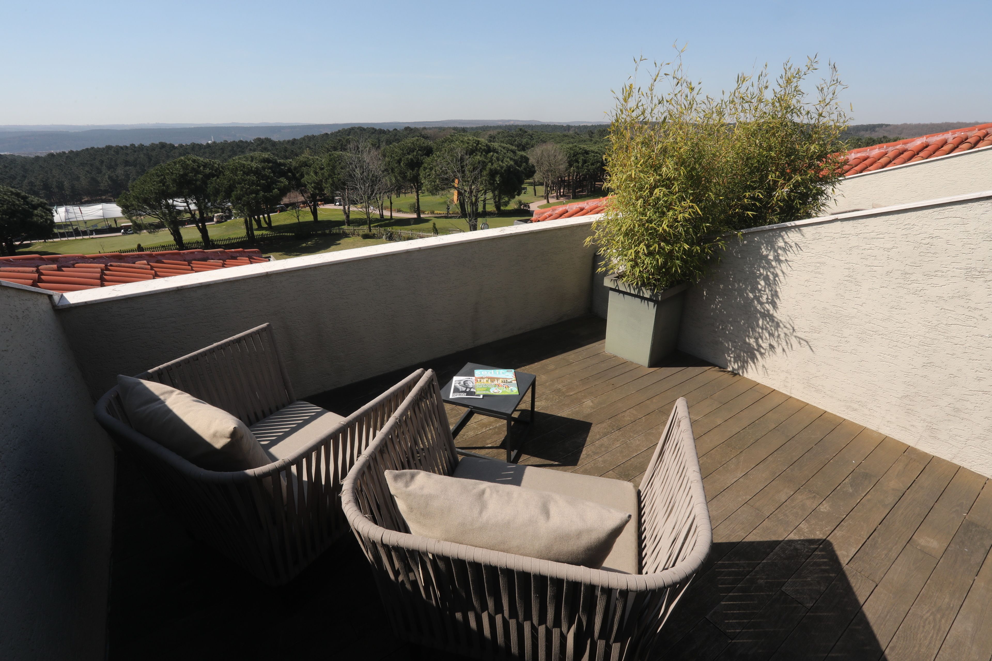 A balcony with wicker furniture and a small table, overlooking a landscaped area with trees and a distant view.