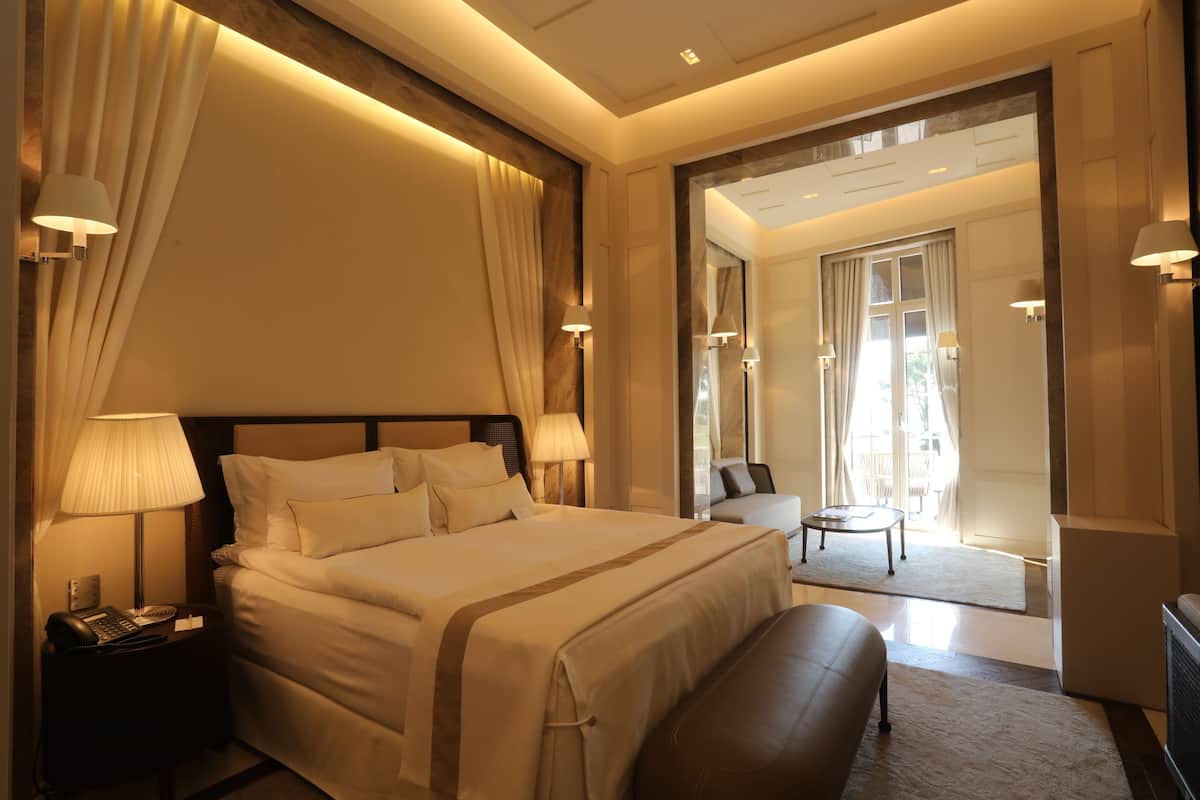 A well-lit hotel room with a large bed, bedside lamps, a desk, and a view of the outdoor area.