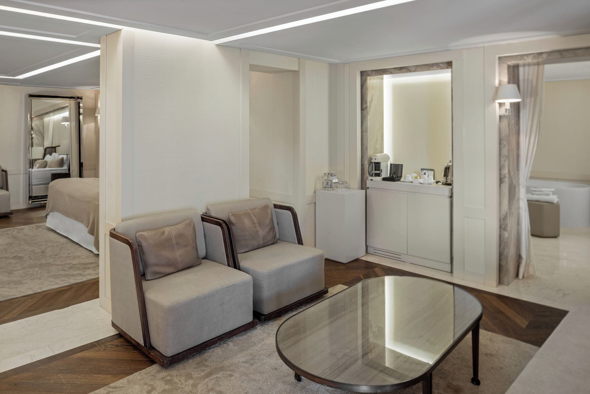 A modern hotel room with a sofa, a glass coffee table, a minibar, and a bathroom with a mirror and curtains.