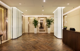Um hall de hotel com piso de madeira em padrão herringbone, um balcão de recepção, plantas em vasos e uma escada que leva aos andares superiores.