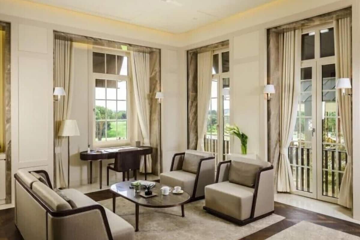 A spacious room with a sofa, armchairs, a coffee table, and a desk. Large windows with curtains provide natural light and views.