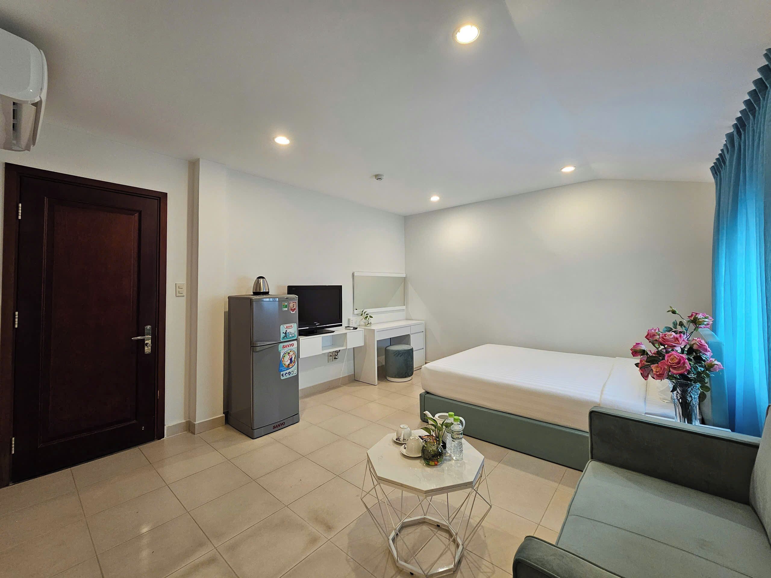 A modern hotel room with a bed, a small desk with a computer, a refrigerator, and a window with curtains.