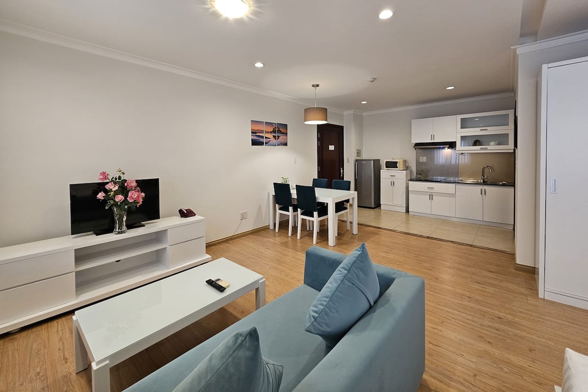 Un salon moderne avec un canapé, un téléviseur et une table basse. On y trouve également un coin repas avec des chaises et une table, ainsi qu’une cuisine équipée de placards et d’appareils électroménagers.