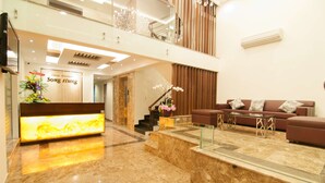 A modern lobby with a reception desk, a staircase, a seating area with a sofa and coffee table, and a decorative plant.