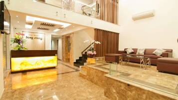 A modern lobby with a reception desk, a staircase, a seating area with a sofa and coffee table, and a decorative plant.