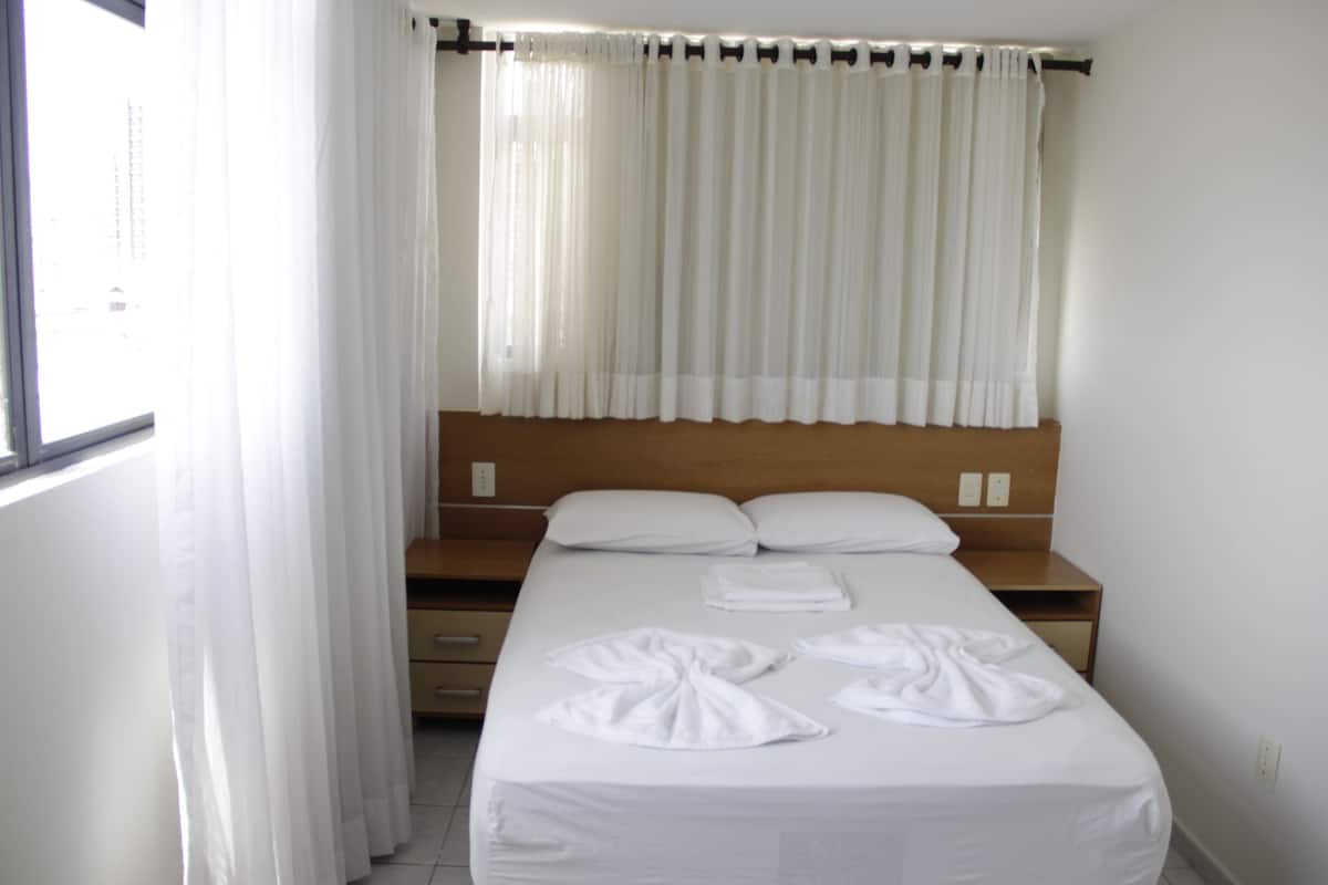 A hotel room with a bed, white curtains, and a wooden headboard.