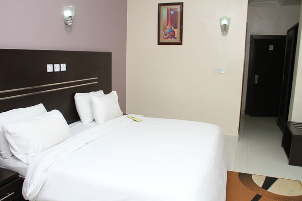 Un cuarto de hotel con una cama grande, cabecera, un cuadro en la pared y una puerta que da a otra habitación.