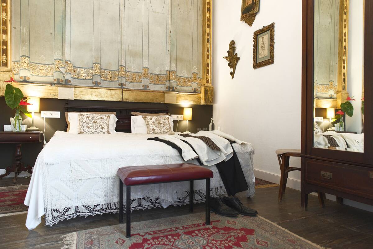 Une chambre bien rangée, avec un grand lit, un banc, une commode et un miroir.