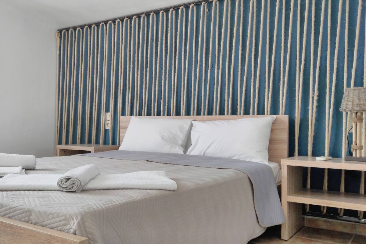 A neatly made bed with white linens and a rolled towel, a bedside table with a lamp, and a vertical rope wall panel.