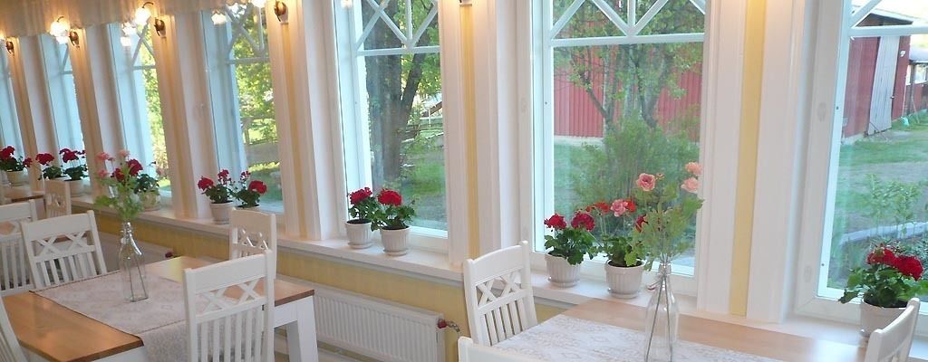 Uma área de refeições com cadeiras e mesas brancas, plantas em vasos com flores vermelhas e grandes janelas com vista para vegetação e um edifício vermelho.